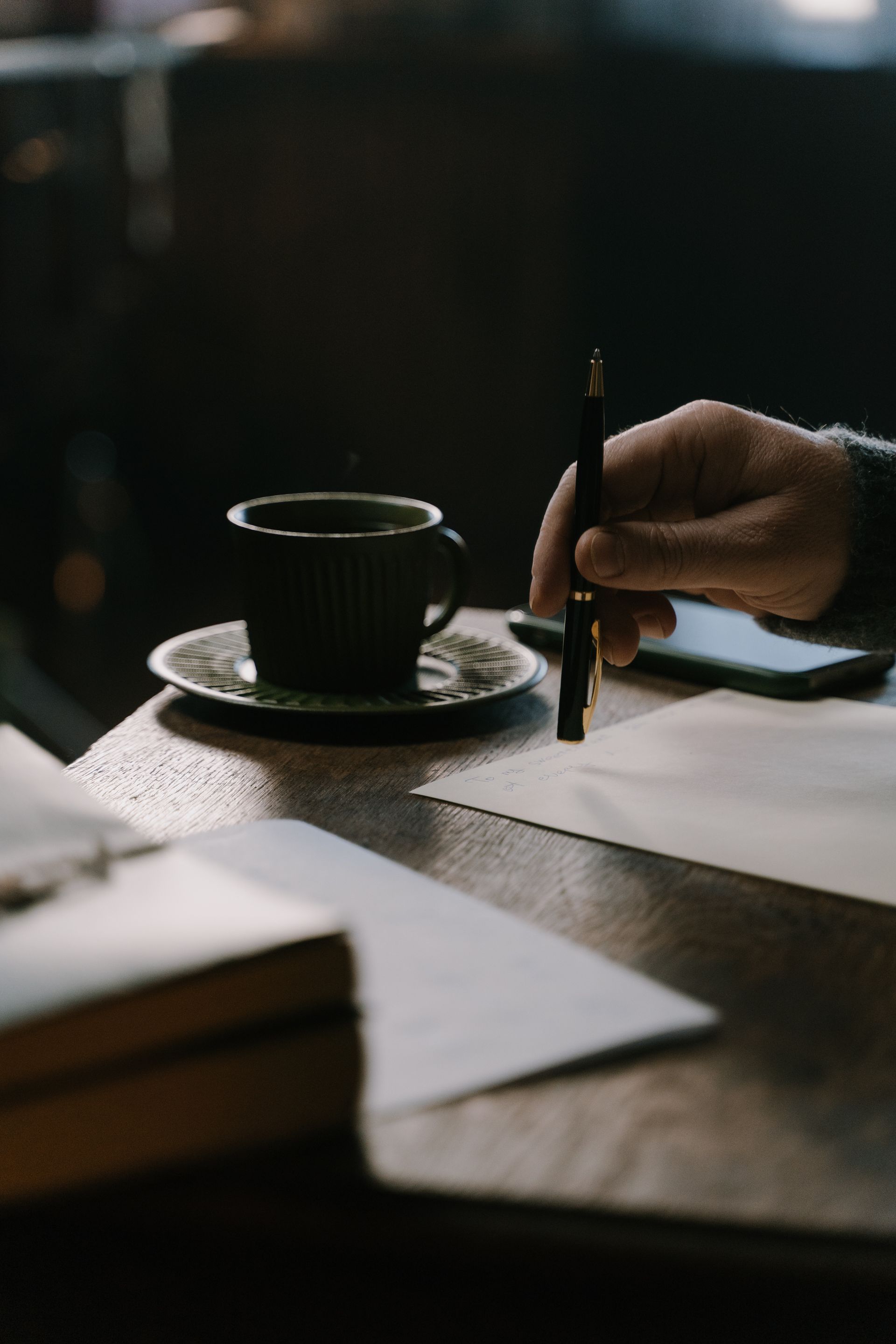 A hand holding a pen over a handwritten letter beside coffee and journals in warm light